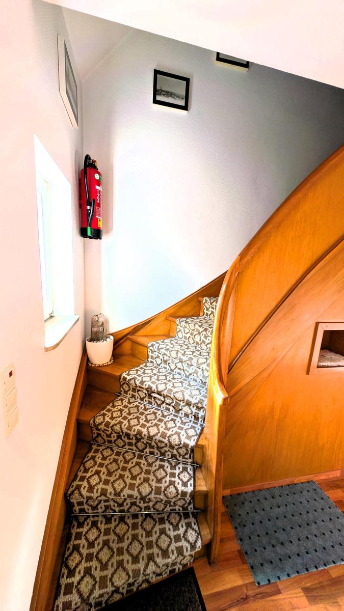 Holzgetreppe mit gemustertem Teppich, Feuerlöscher an der Wand und kleines Fenster.