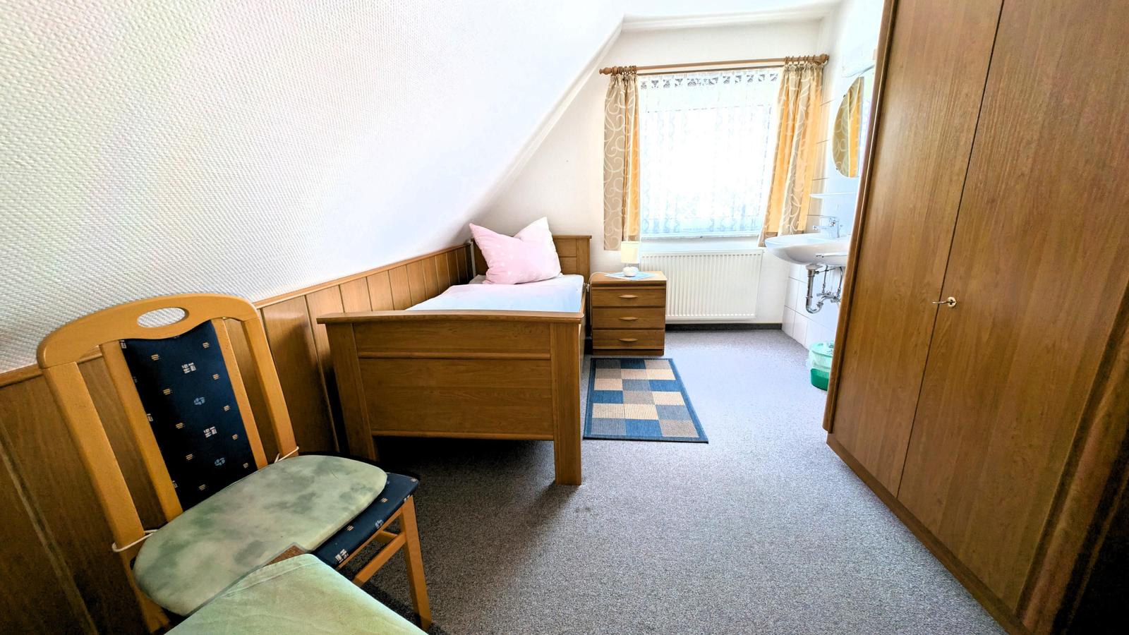 Schlafzimmer mit Bett, Stuhl, Schrank und Fenster mit Vorhängen.