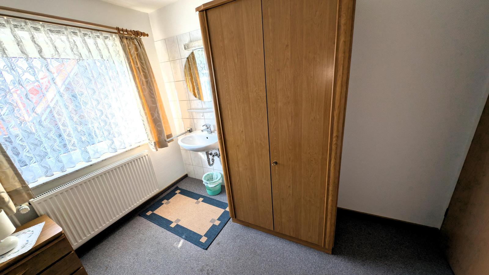Zimmer mit Kleiderschrank, Waschbecken und Fenster mit Vorhängen.