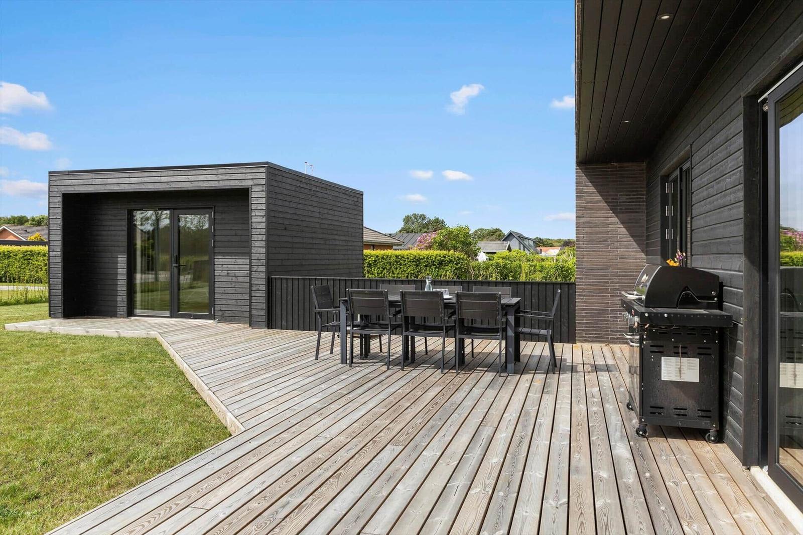 Terrasse mit Tisch, Stühlen und Grill. Hintergrund: modernes Haus mit Holzverkleidung.