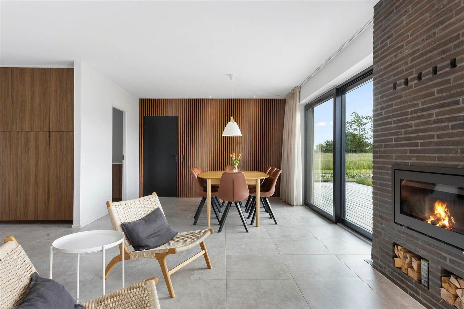 Wohn- und Essbereich mit Kamin, Holztisch und Sitzgelegenheiten. Große Fenster mit Blick auf die Außenterrasse.