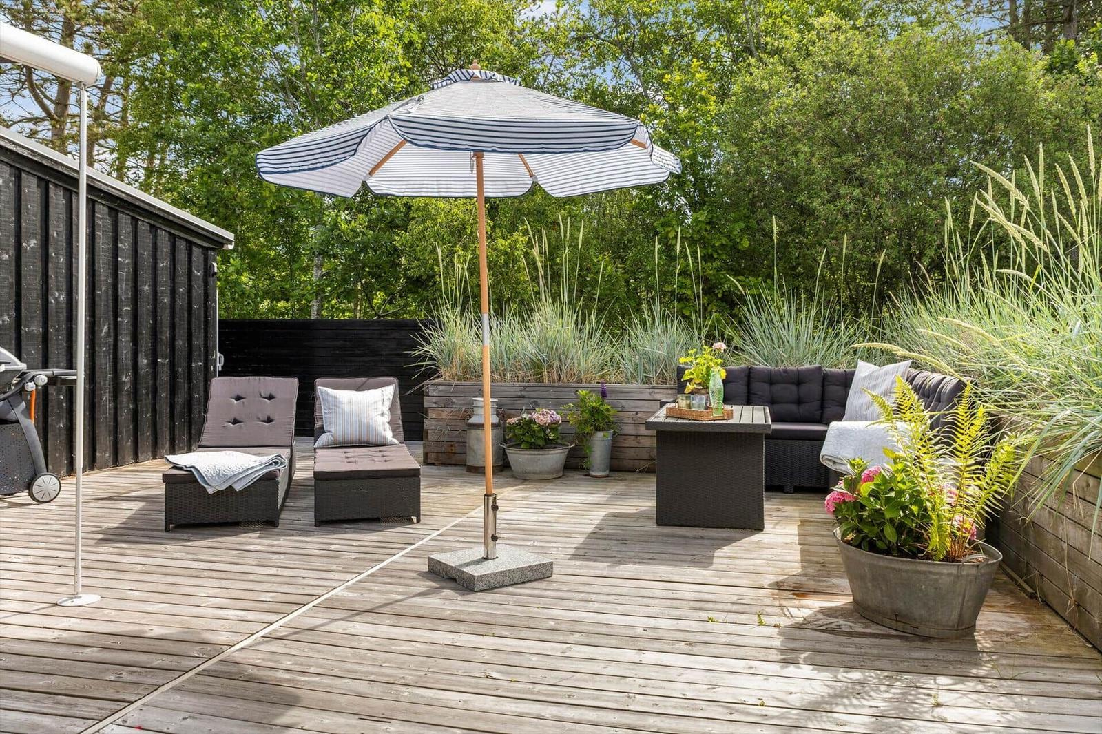 Deck with lounge furniture, umbrella, and plants. Wooden deck and trees in background.
