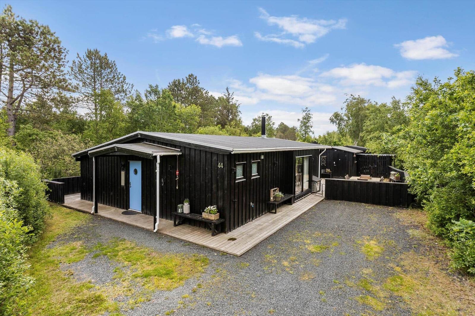 Black wooden house with blue door, deck, and garden. Surrounded by trees and grass.