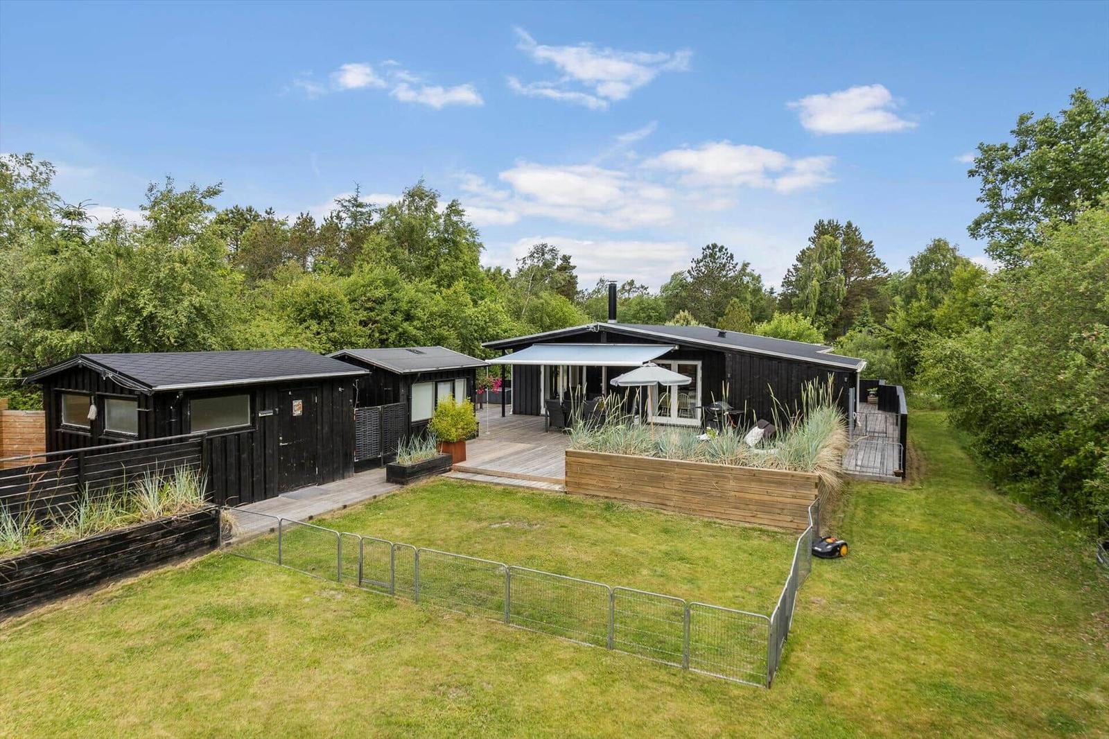 Black wooden cabin with terrace and garden. Surrounded by trees under blue sky.