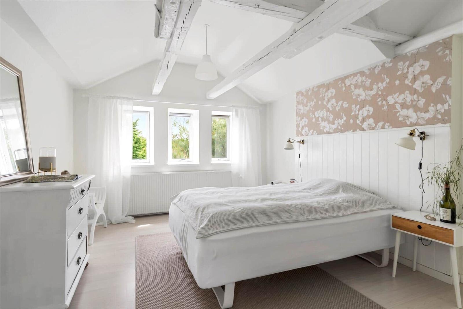 Schlafzimmer mit weißem Bett, Holzbalken und floraler Wandverkleidung.