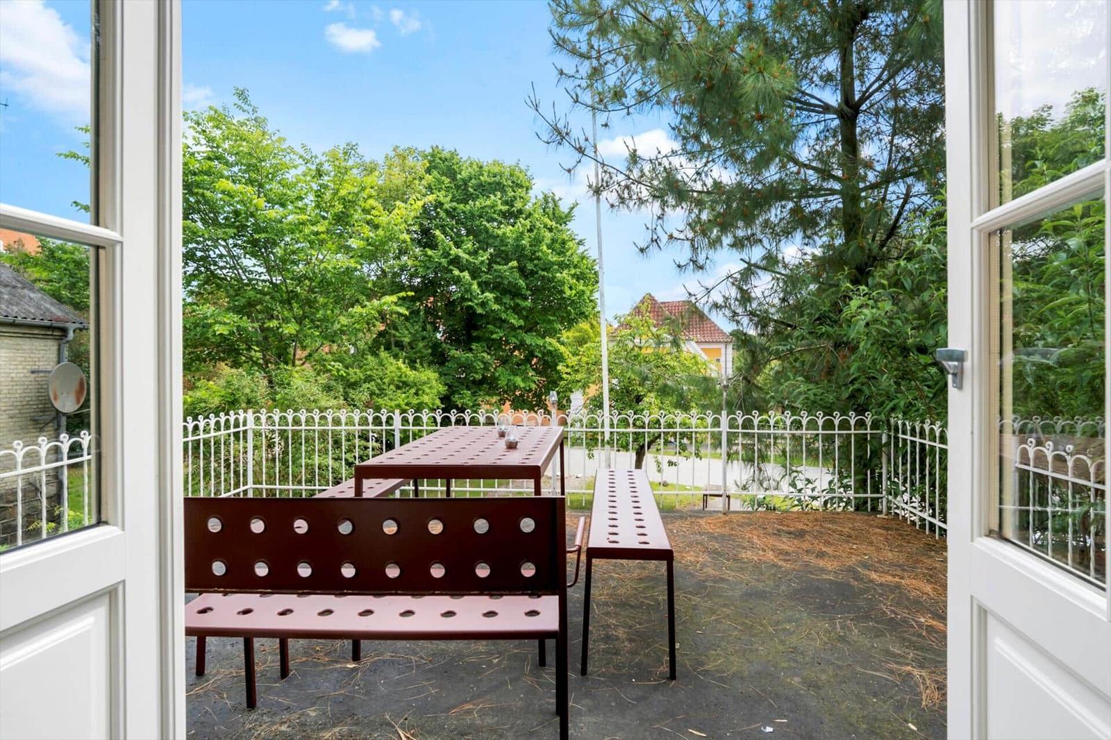 Terrasse mit Tisch und Bänken, umgeben von grünen Bäumen und einem weißen Zaun.