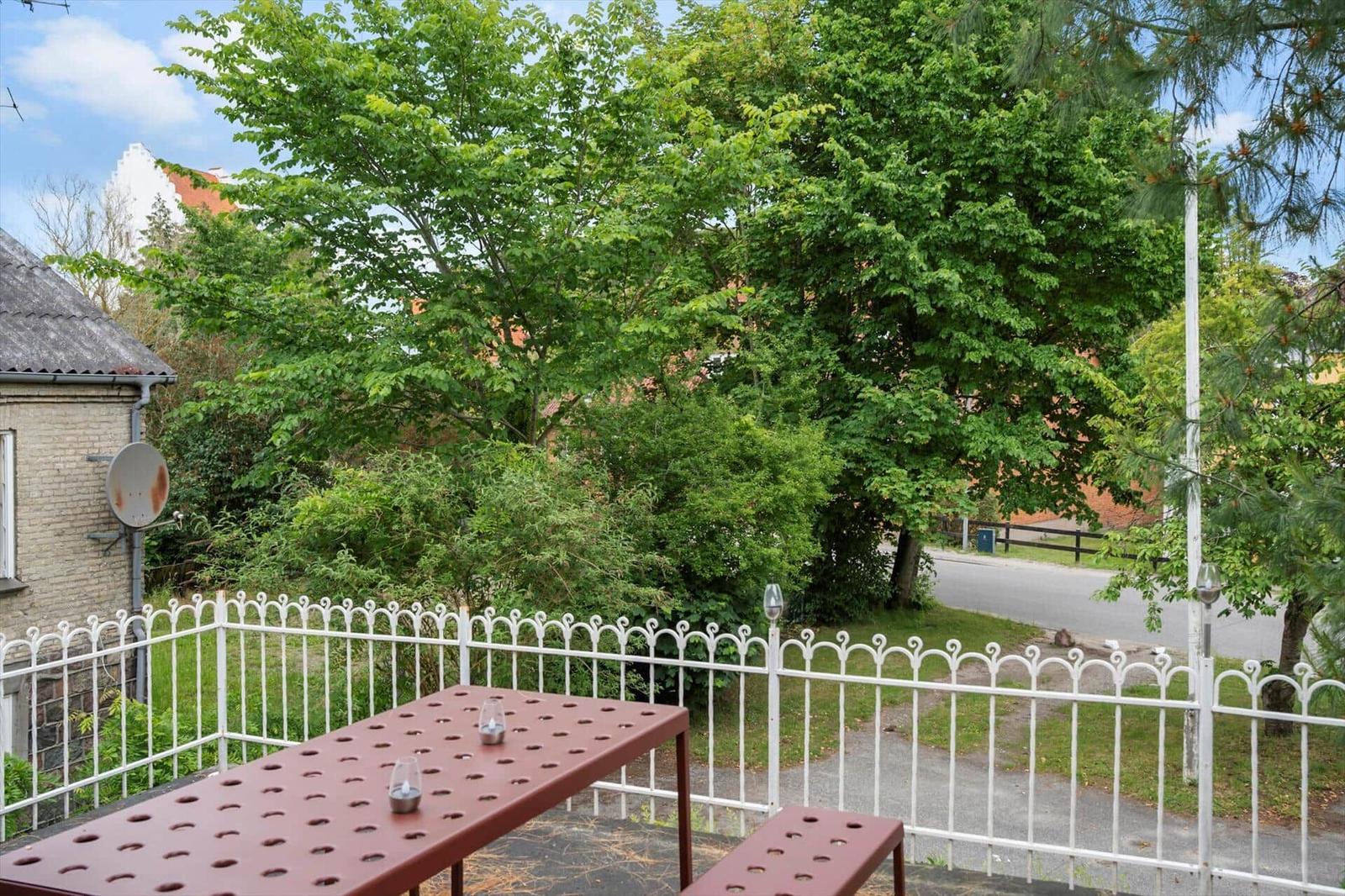Terrasse mit Tisch und Bänken, umgeben von weißen Metallgitter und grünen Bäumen.