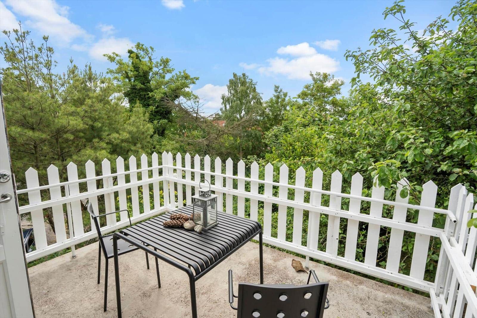Terrasse mit weißem Zaun und schwarzem Tischset. Blick in grüne Bäume und blauen Himmel.