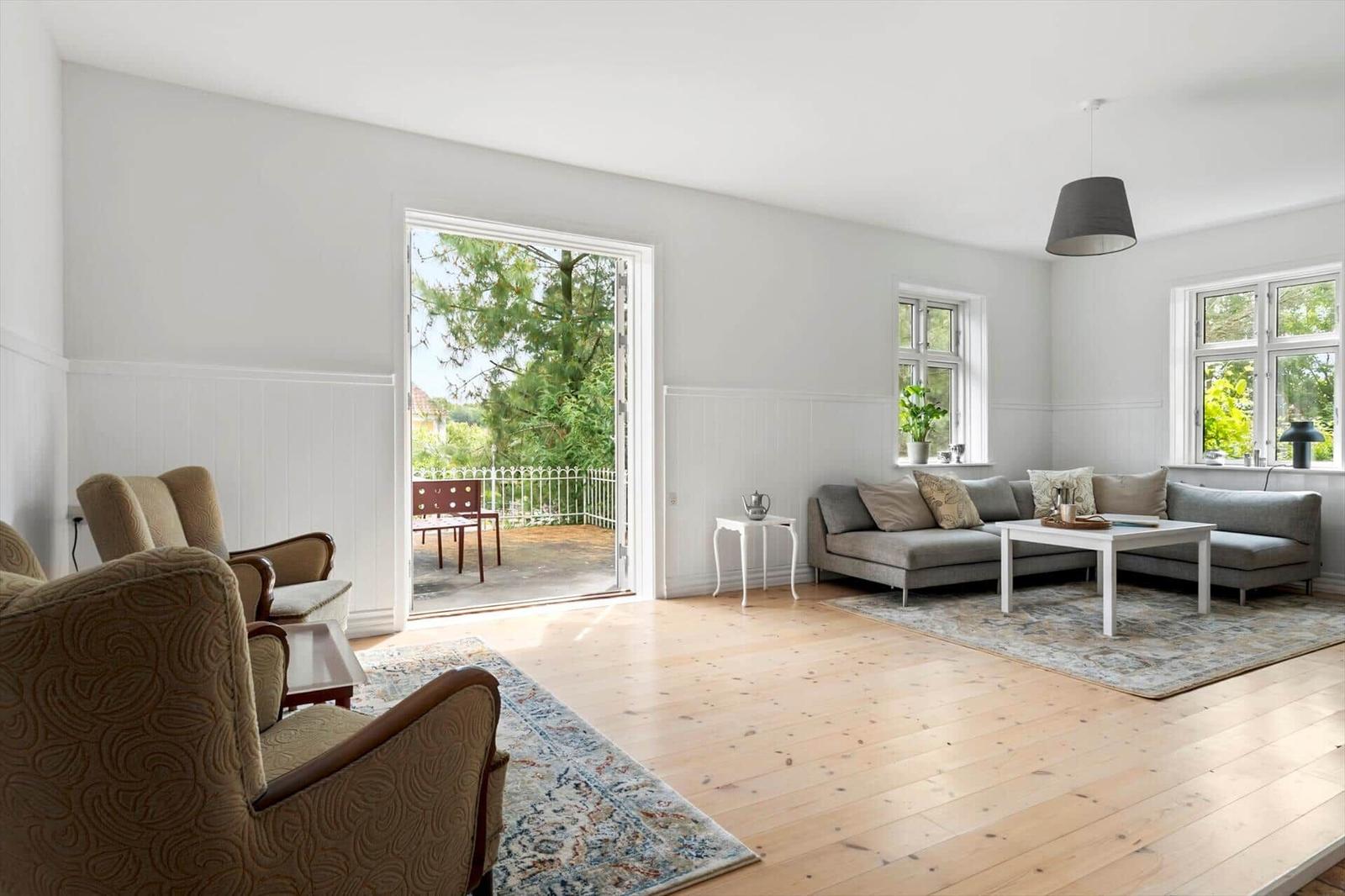 Wohnzimmer mit Sofa, Sessel und Terrasse. Holzboden und Fenster mit Blick auf Grün.