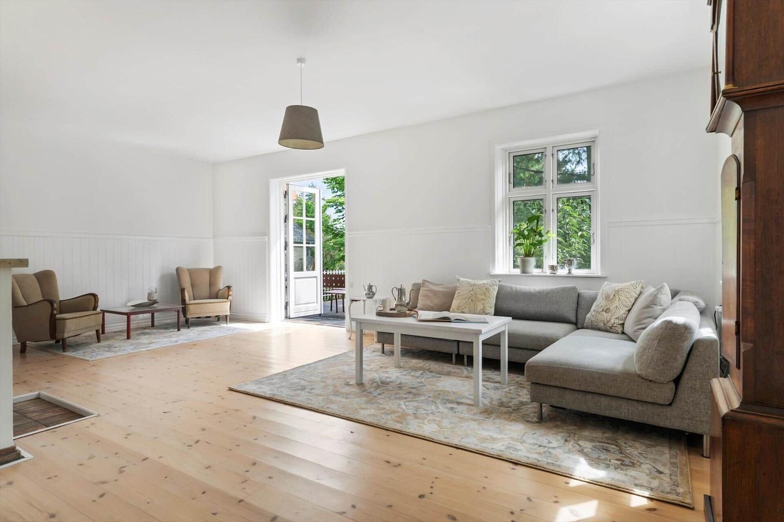 Wohnzimmer mit Sofa, zwei Sesseln, Tisch und Fenster. Holzboden und Tür zum Garten.