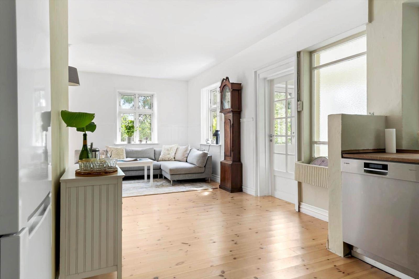 Wohnzimmer mit Sofa, Holzboden und Uhr. Küche mit Spülmaschine sichtbar.