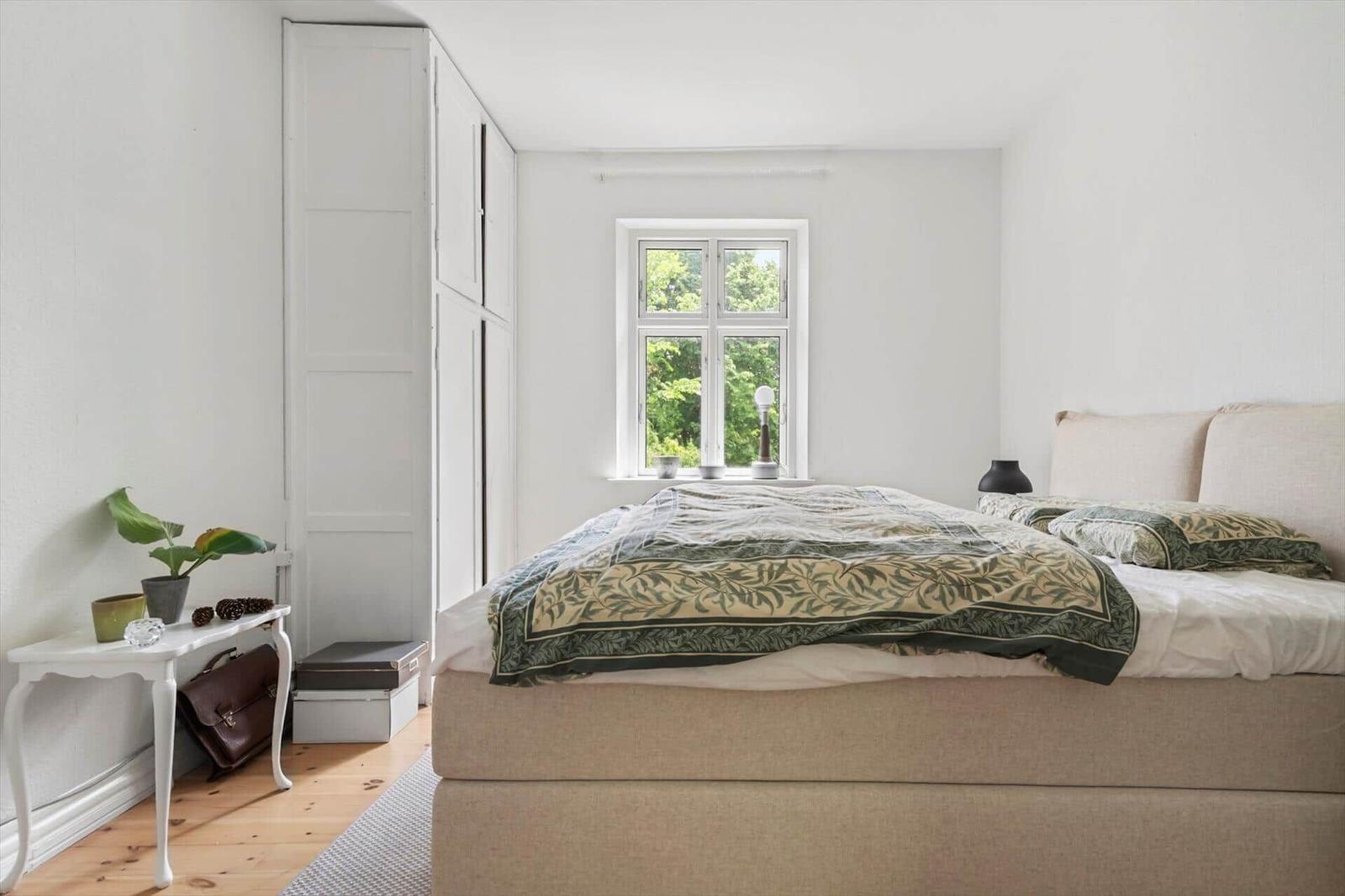 Schlafzimmer mit Bett, Fenster und Kleiderschrank. Holzboden und weiße Wände.