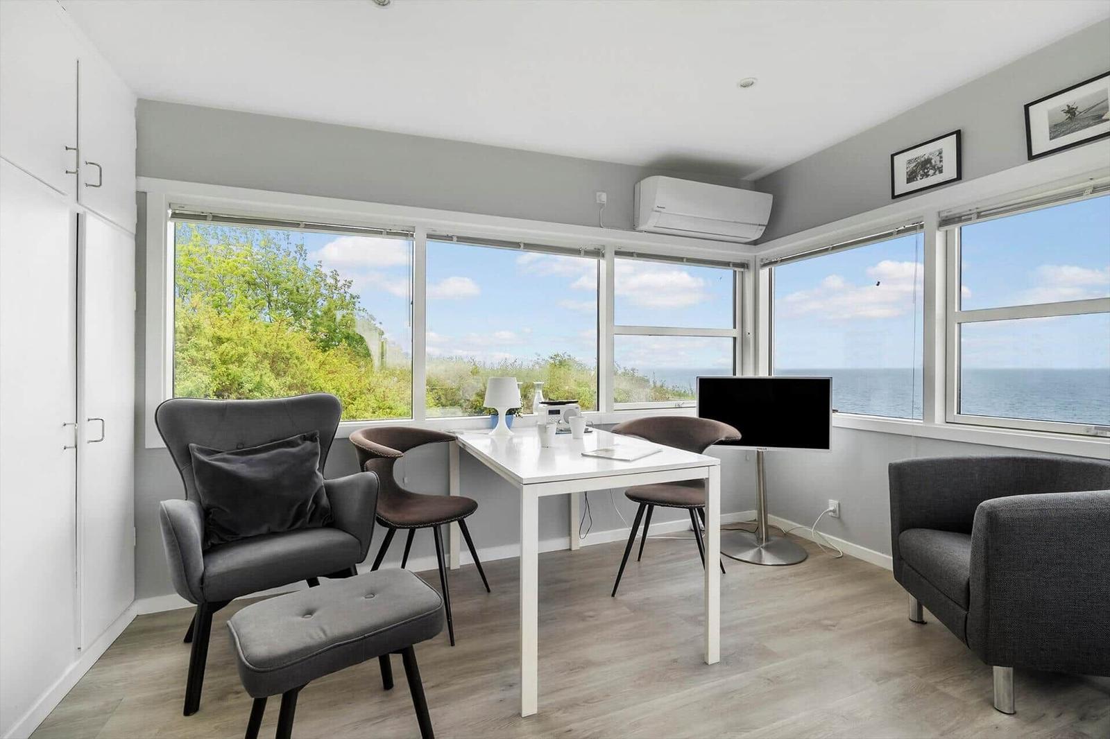 Ein Raum mit Tisch, Stühlen und Fernseher. Große Fenster bieten Blick auf Meer und Bäume.