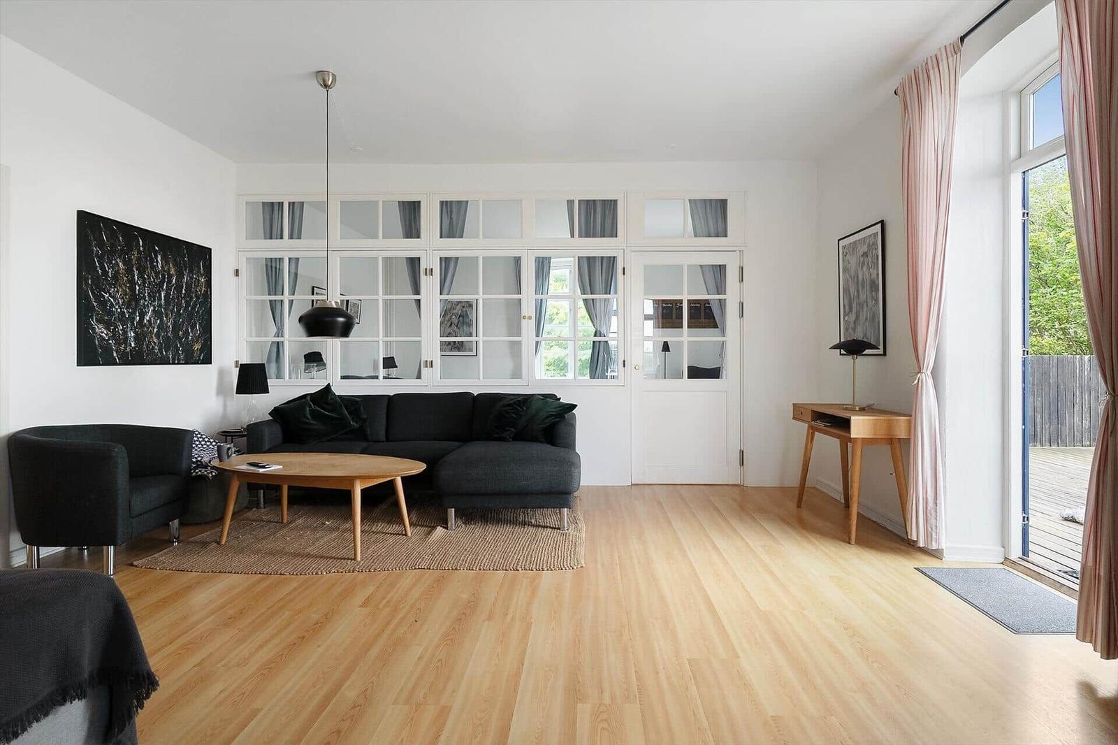 Wohnzimmer mit Sofa, Couchtisch und Holzboden. Große Fenster und Tür führen nach draußen.