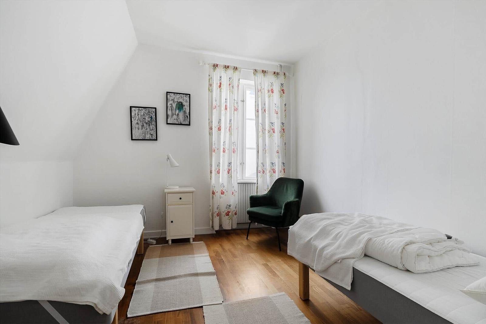 Zwei Betten, ein grüner Sessel, Fenster mit floralen Vorhängen und Holzboden.