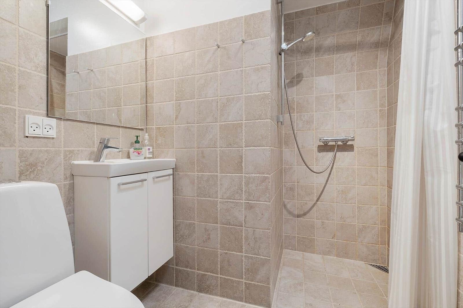 Bad mit Dusche, Waschbecken und Toilette. Fliesenwand und Spiegel sichtbar.