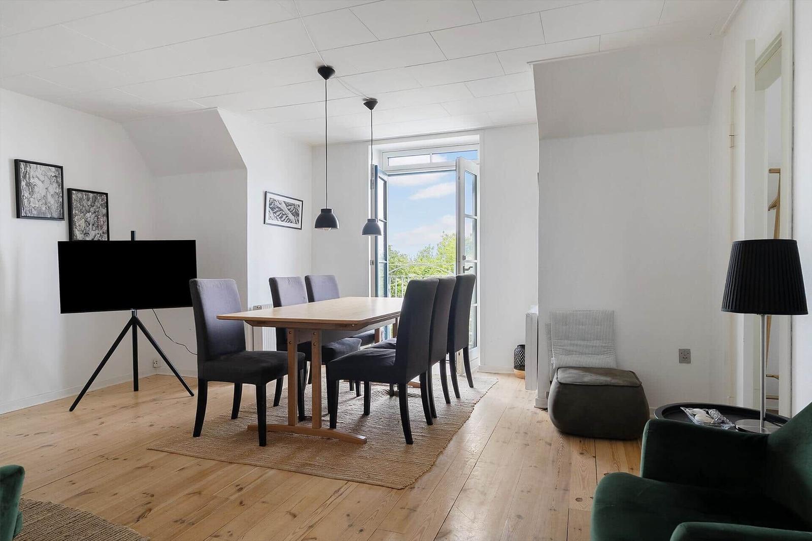 Essbereich mit Tisch, Stühlen und TV. Tür führt auf Balkon. Holzboden und weiße Wände.