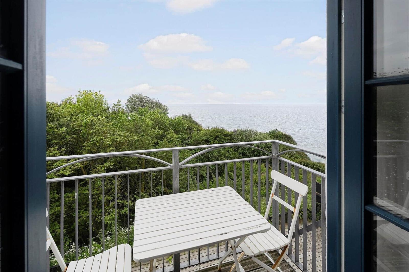 Balkon mit weißem Tisch und Stühlen, Blick auf Meer und grüne Bäume.