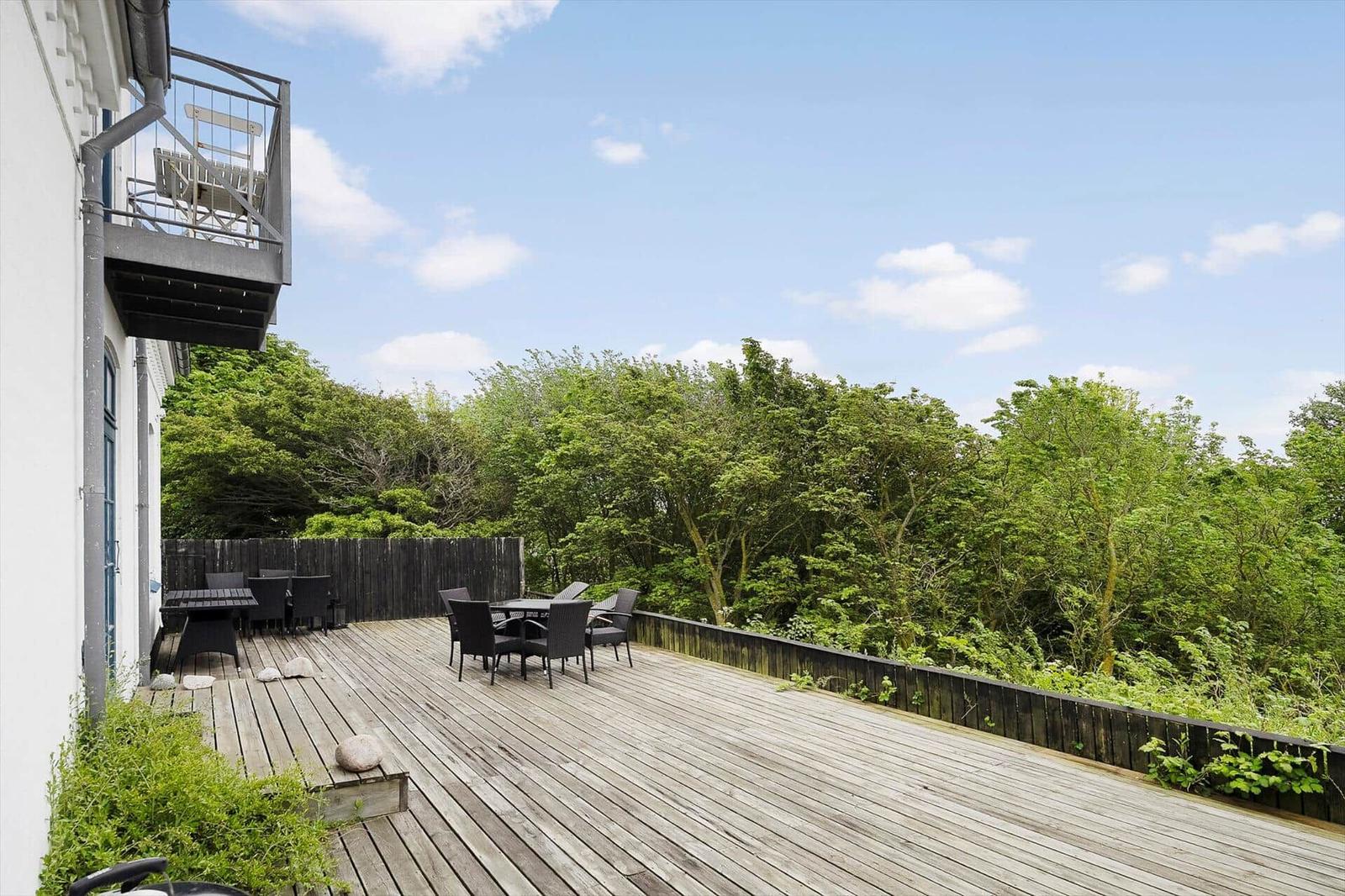 Holzterrasse mit Sitzgruppe und Blick auf grüne Bäume. Hinterhof mit Holzzaun und Balkon.