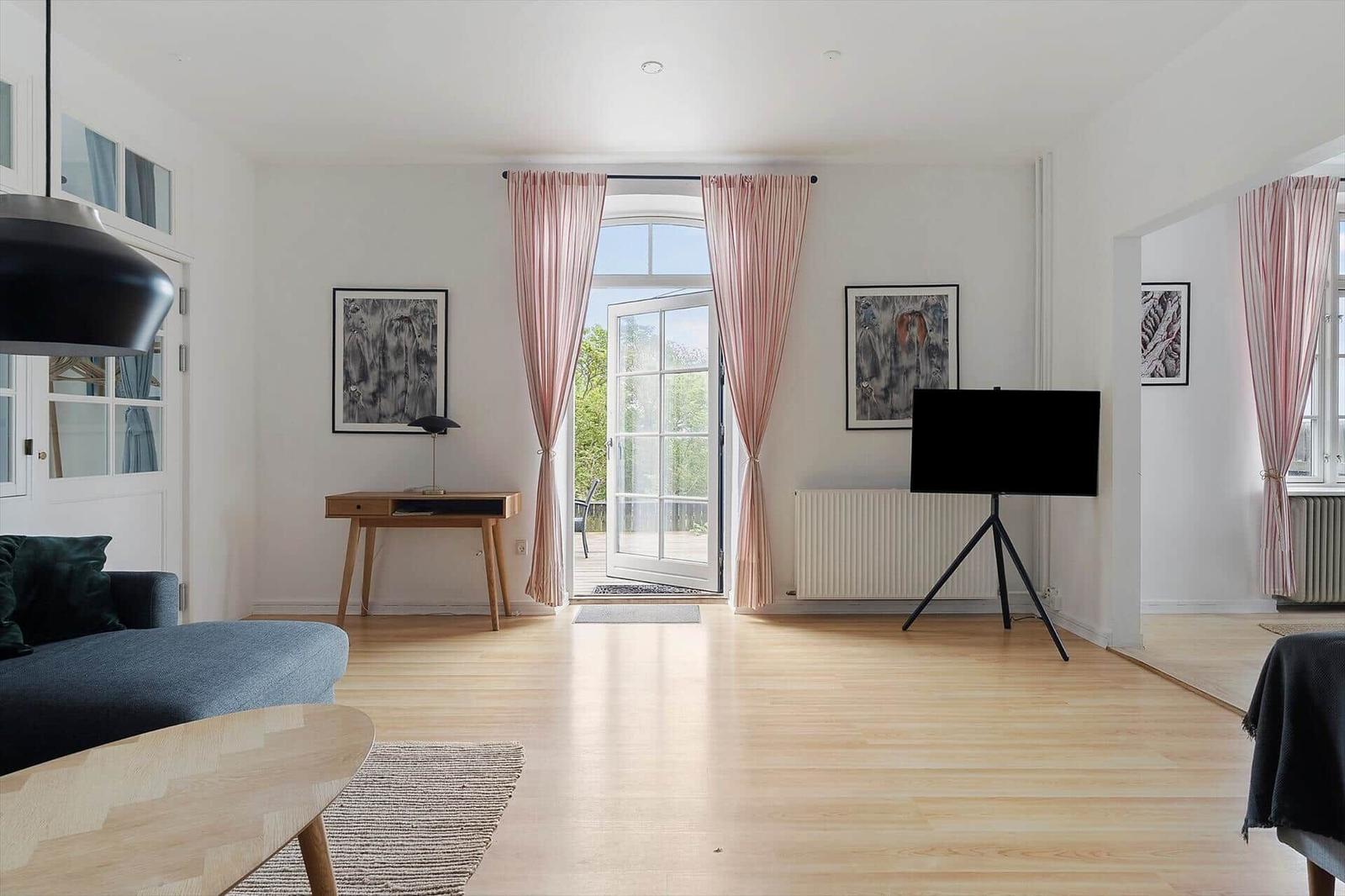 Wohnzimmer mit Holzboden, Sofa, Fernseher auf Stativ und Fenster mit rosa Vorhängen.