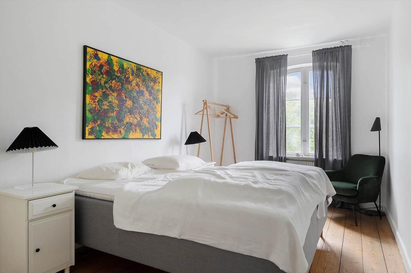 Schlafzimmer mit Bett, Nachttisch, Stuhl und Fenster mit Vorhängen.