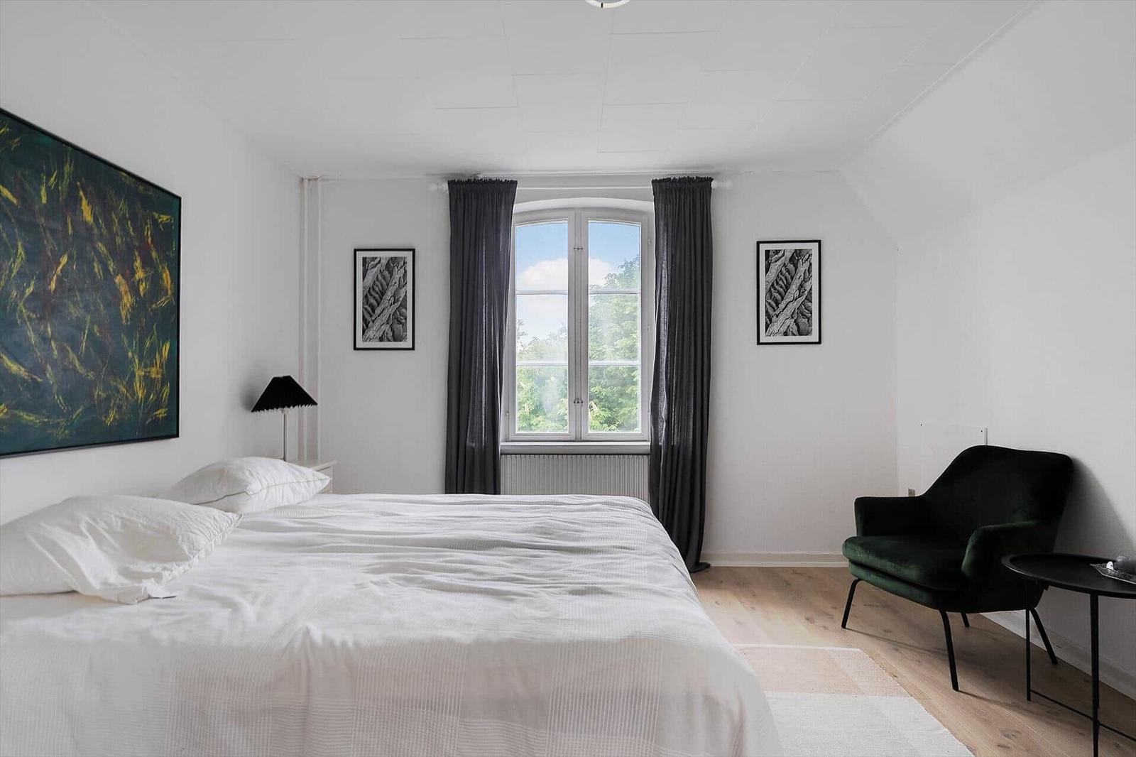 Schlafzimmer mit großem Bett, Fenster mit Blick auf Grün, dunkler Sessel und Wandbild.