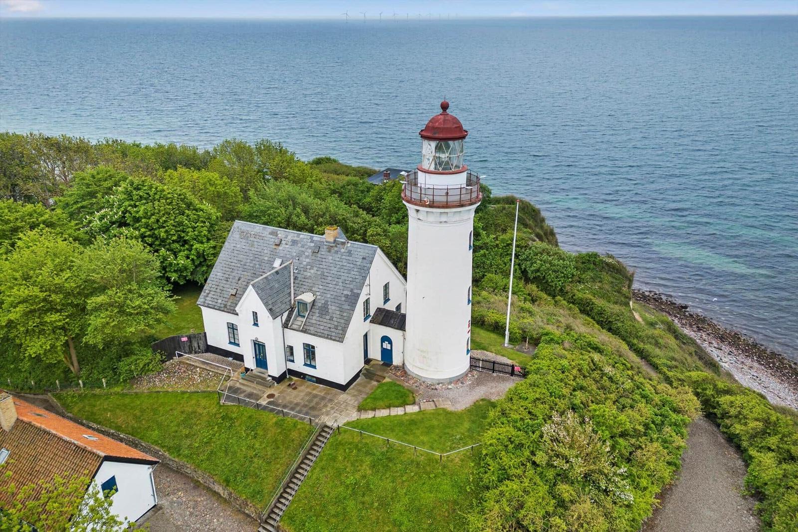 Weißes Haus und Leuchtturm auf grüner Halbinsel am Meer mit Windkraftanlagen im Hintergrund.