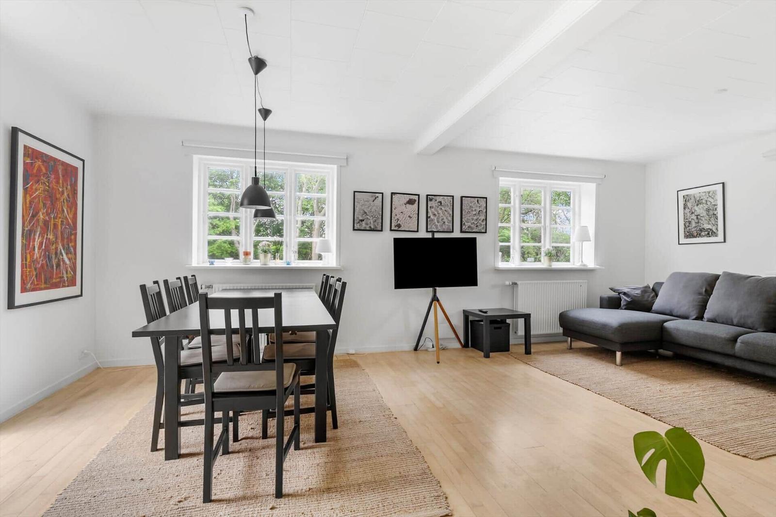 Wohn- und Essbereich mit Esstisch, Sofa und TV. Holzboden und große Fenster.