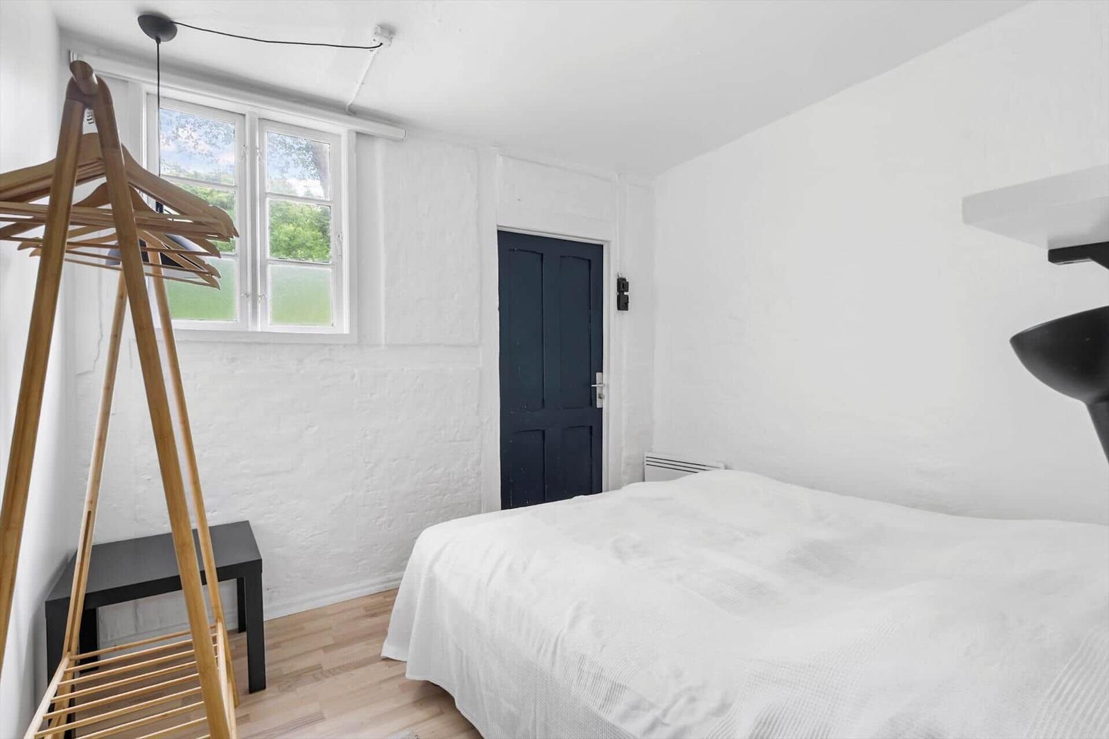Schlafzimmer mit weißer Wand, Bett, Fenster und Holzständer.