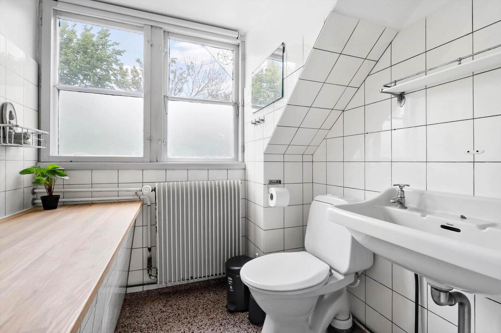 Badezimmer mit Toilette, Waschbecken und Fenster. Weiße Fliesen und Holzbank.