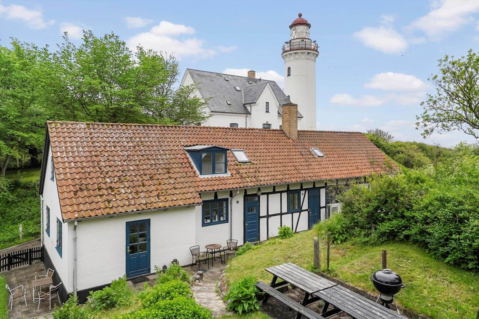 Wohnhaus mit rotem Ziegeldach und blauen Türen. Im Hintergrund Leuchtturm. Grünanlagen und Grillplatz.