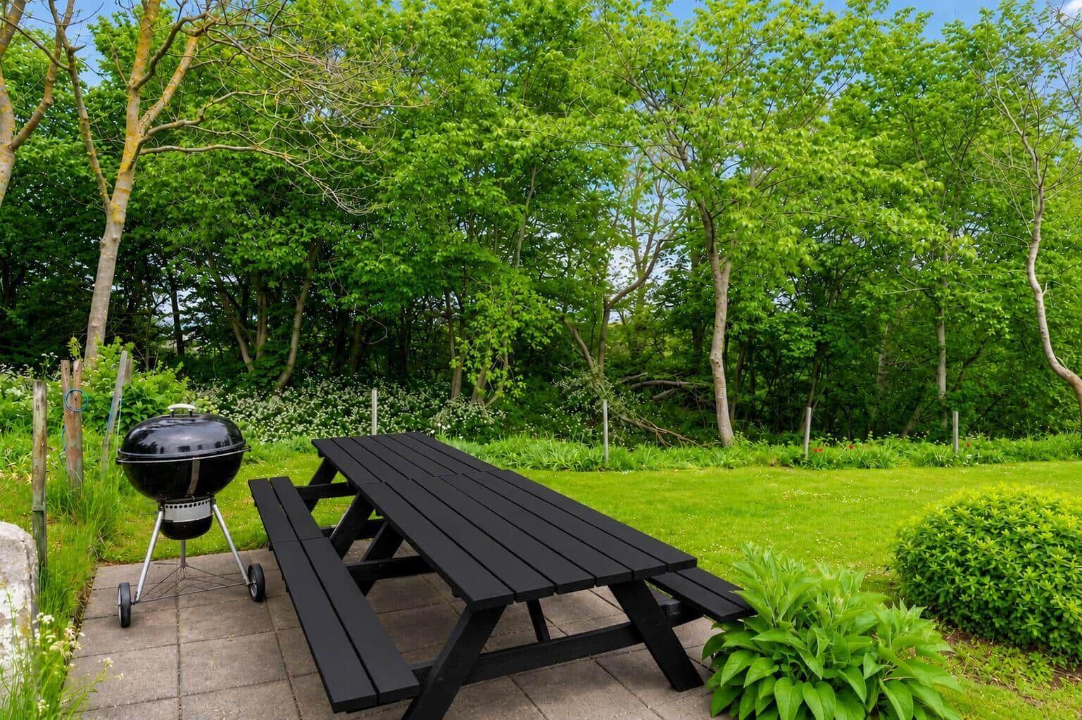 Schwarzer Picknicktisch und Grill auf Terrasse mit Grünfläche und Bäumen im Hintergrund.
