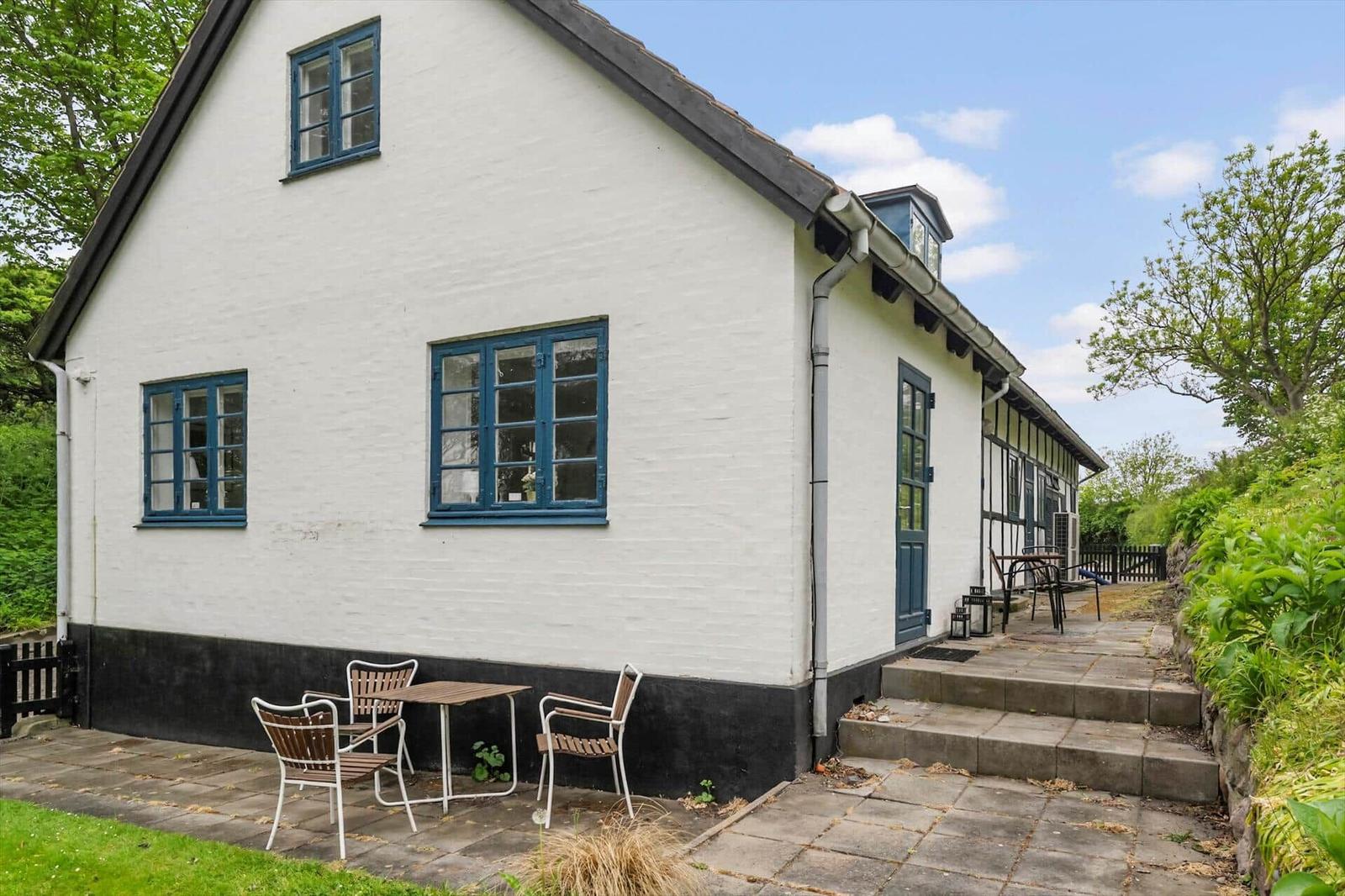 Weiße Villa mit blauen Fenstern und Terrasse. Garten und Holzhaus im Hintergrund.