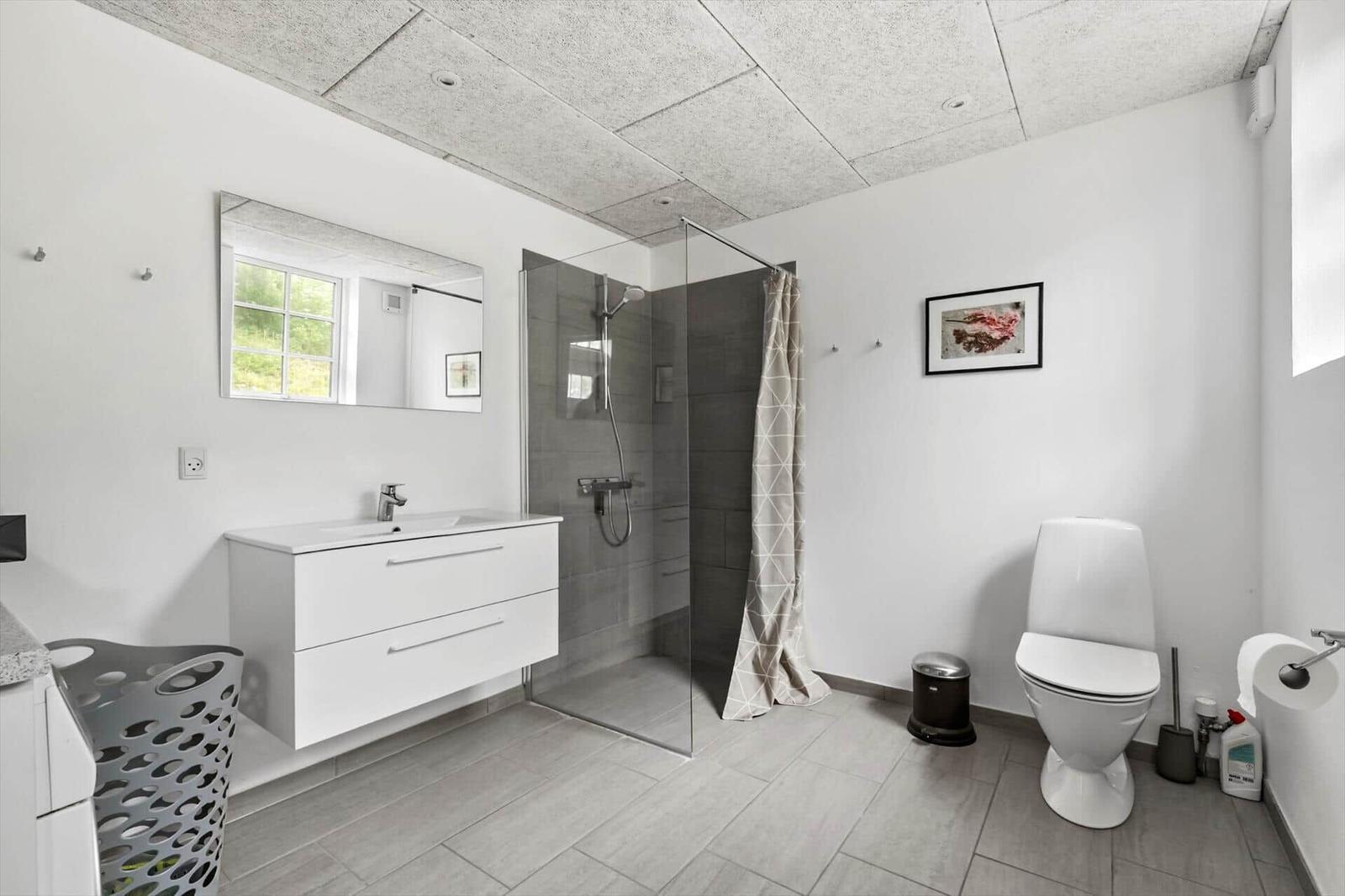 Badezimmer mit Dusche, Waschbecken und Toilette. Weiße Wände, graue Fliesen und Fenster.