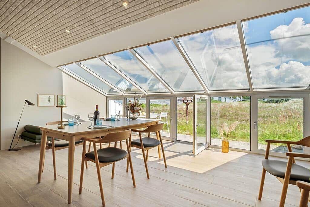 Essbereich mit Tisch, Stühlen und Glasdach. Blick auf Grünflächen und Himmel.