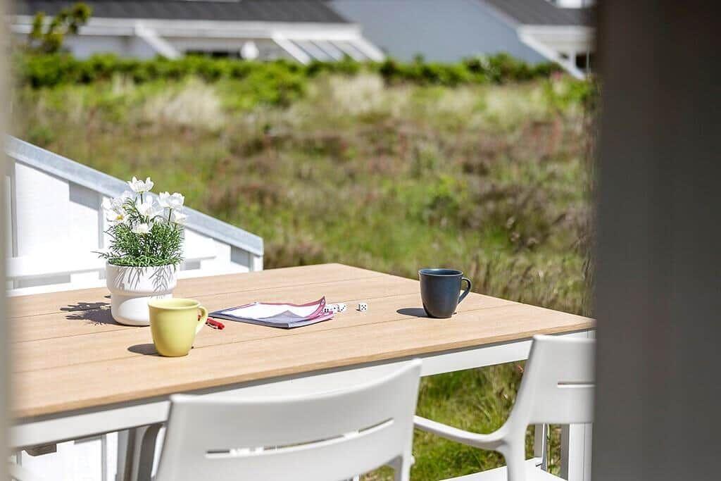 Holztisch mit zwei Tassen, Pflanze und Würfeln auf einer Terrasse mit Grünfläche im Hintergrund.