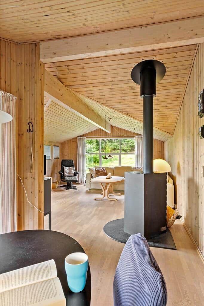 Wohnraum mit Holzwand, Kaminofen und Sitzgelegenheiten. Durchgang zu weiterem Raum mit Fenster.
