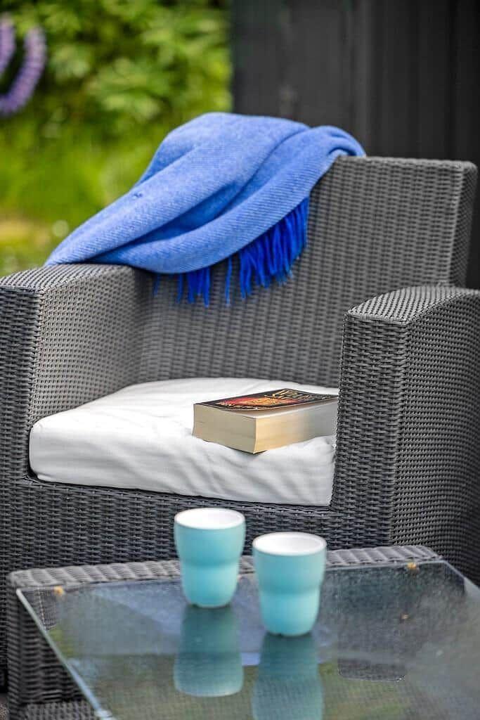 Gartenstuhl mit Kissen, Decke und Buch. Auf Tisch stehen zwei Tassen.