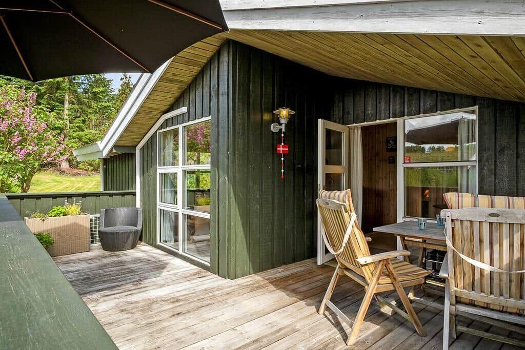 Terrasse mit Holzboden, Stuhl, Tisch und Sonnenschirm vor einem Haus mit grünem Holzverkleidung.