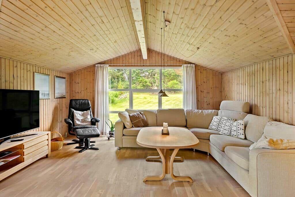 Wohnzimmer mit Holzwand, Sofa, Esstisch und Fernseher. Große Fenster mit Blick auf Grünflächen.