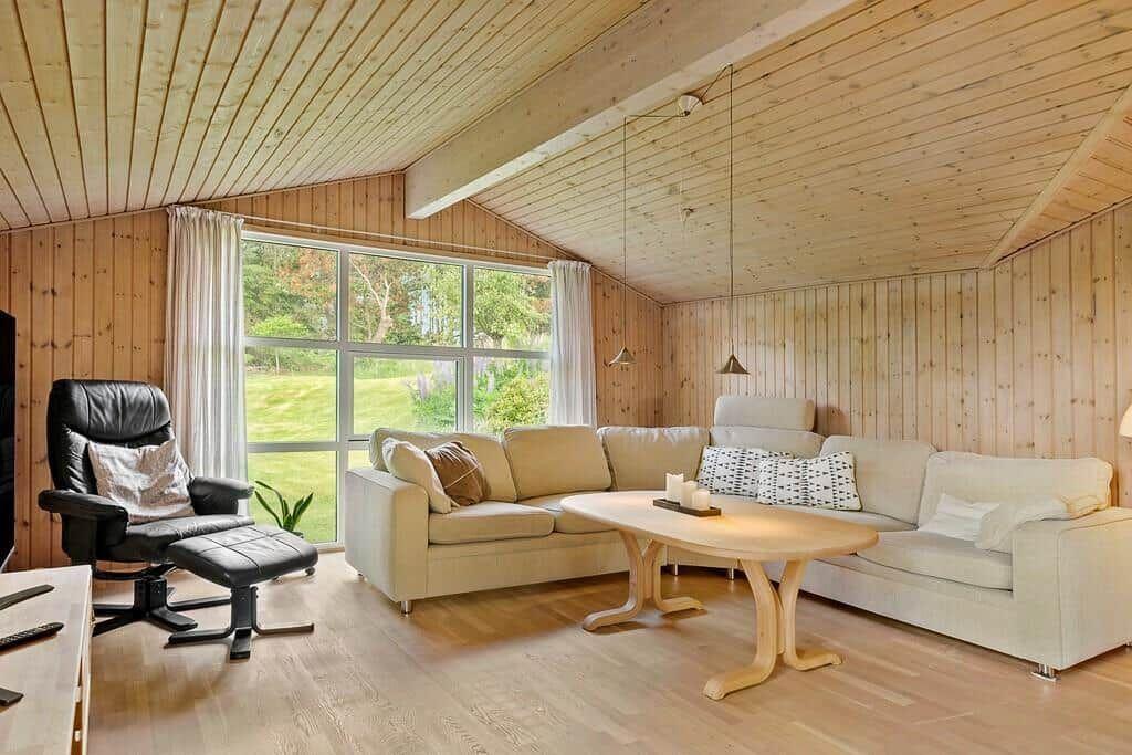 Wohnzimmer mit Holzwand, Sofa, Esstisch und Sessel vor großem Fenster.