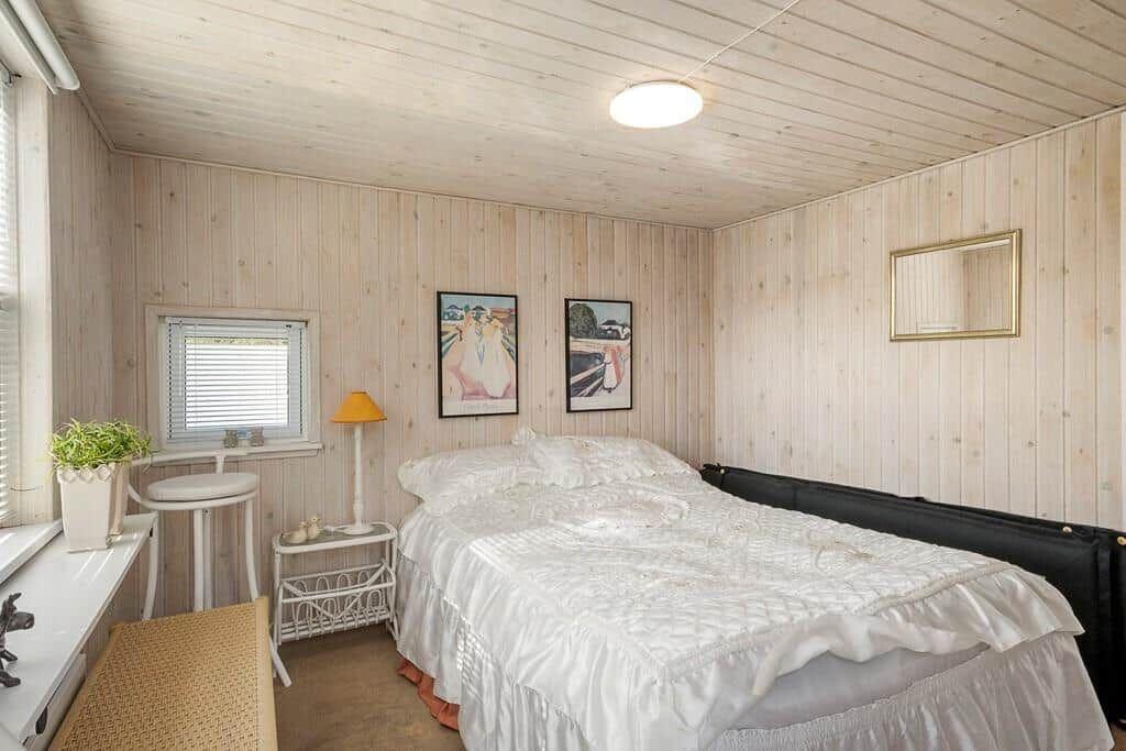 Schlafzimmer mit Holzwandverkleidung, Bett mit weißer Bettwäsche und Fenster mit Jalousie.