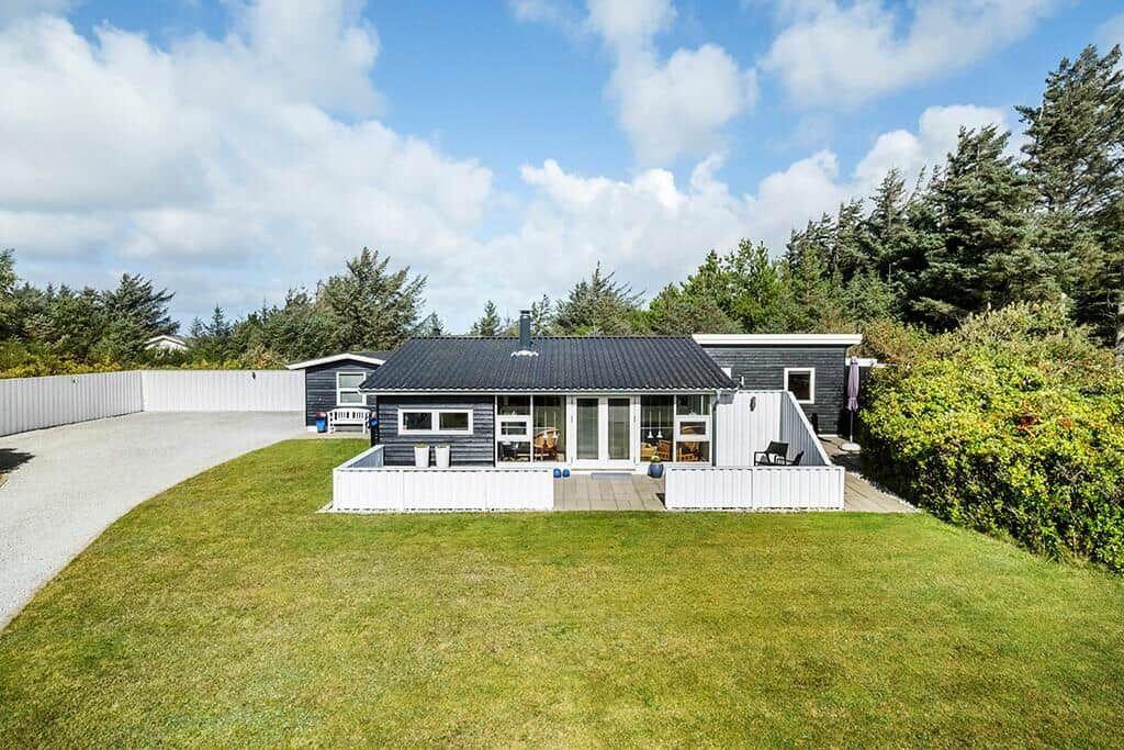 Ein Haus mit dunklem Dach und weißem Zaun auf grünem Rasen. Hintergrund: Bäume und blauer Himmel.