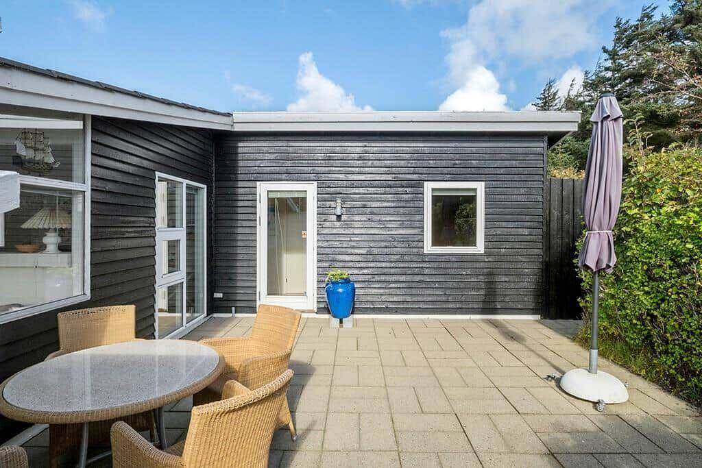 Terrasse mit Tisch, Stühlen und Sonnenschirm. Haus mit dunkler Holzfassade und weißen Fenstern.