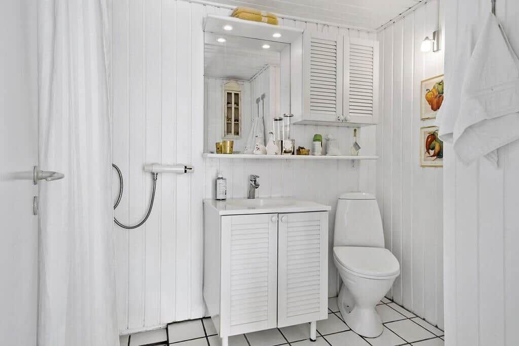 Badezimmer mit Dusche, Waschbecken und Toilette. Weiße Holzwandverkleidung und Fliesenboden.