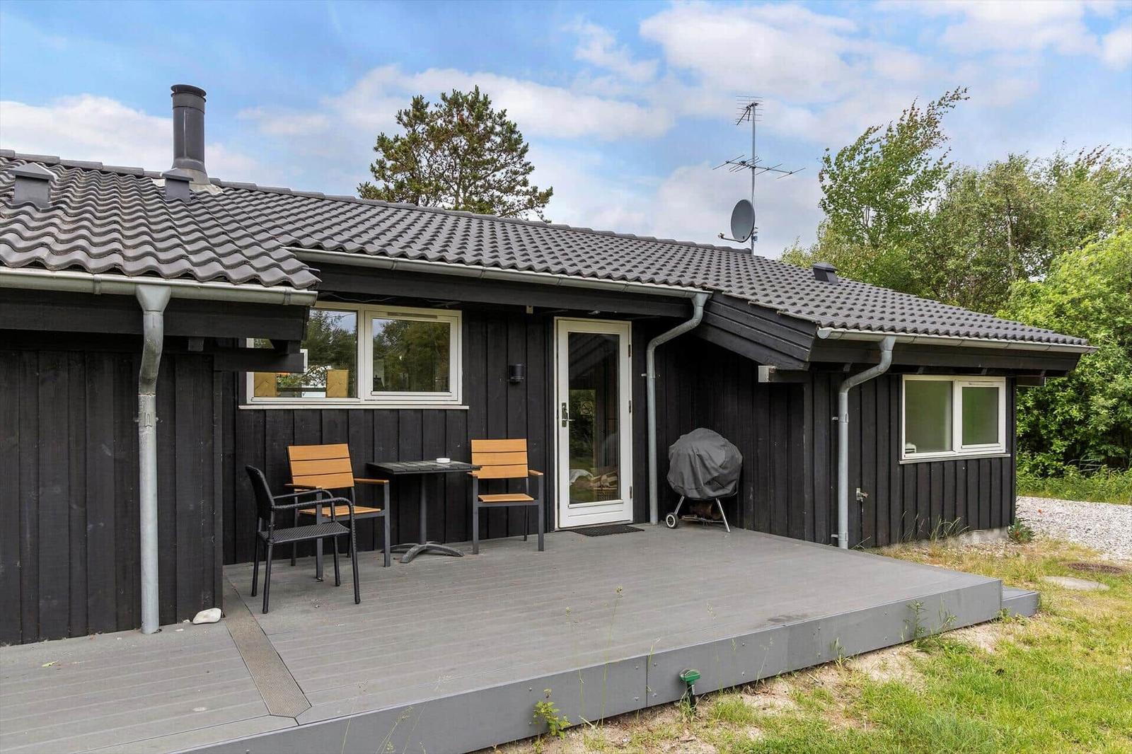 A house with a terrace, table, chairs, and grill. Surrounded by trees and grass.