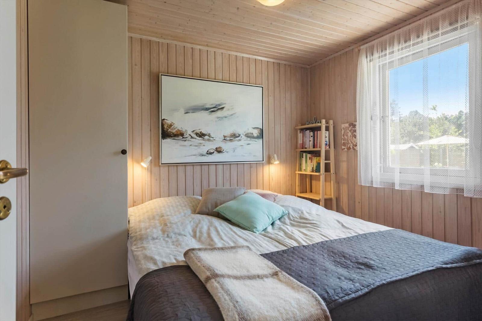Schlafzimmer mit Bett, Wandbild und Bücherregal. Holzwand und Fenster mit Blick nach draußen.