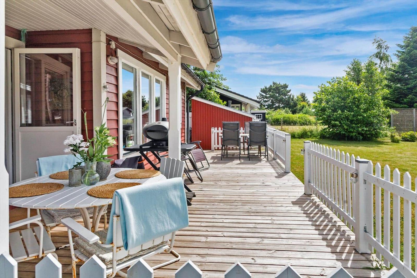 Terrasse mit Tisch, Stühlen und Grill. Roter Holzaufbau, weißer Zaun und Garten.
