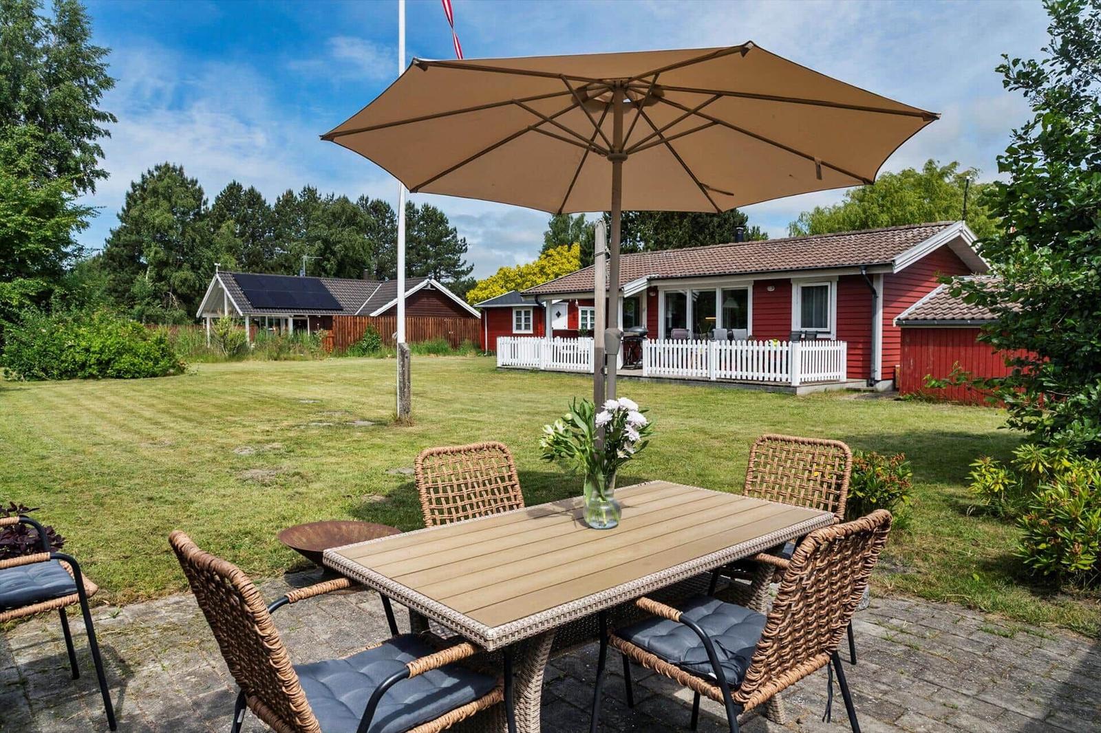 Terrasse mit Tisch, Stühlen und Sonnenschirm. Roter Holzaufbau im Hintergrund.