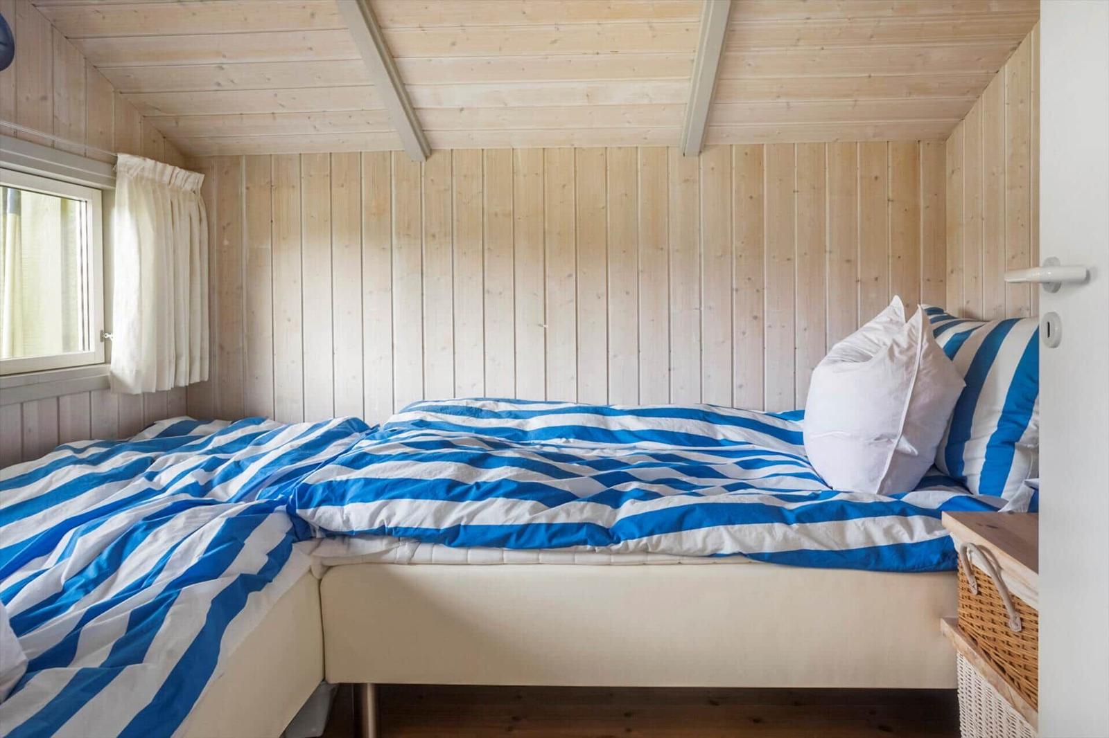 Schlafzimmer mit Holzwand, Bett mit blau-weißem Bettzeug und Fenster mit Vorhang.
